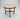 A round outdoor accent table with a mosaic tile top in beige and white, supported by a dark brown metal frame with four legs and a central stretcher bar, set on a concrete patio.